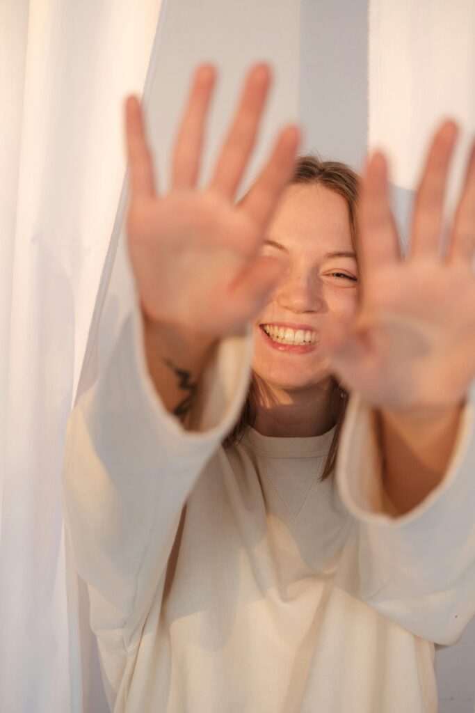 Smiling young woman raises raises to the camera through sheer curtains in bright, sun-lit room